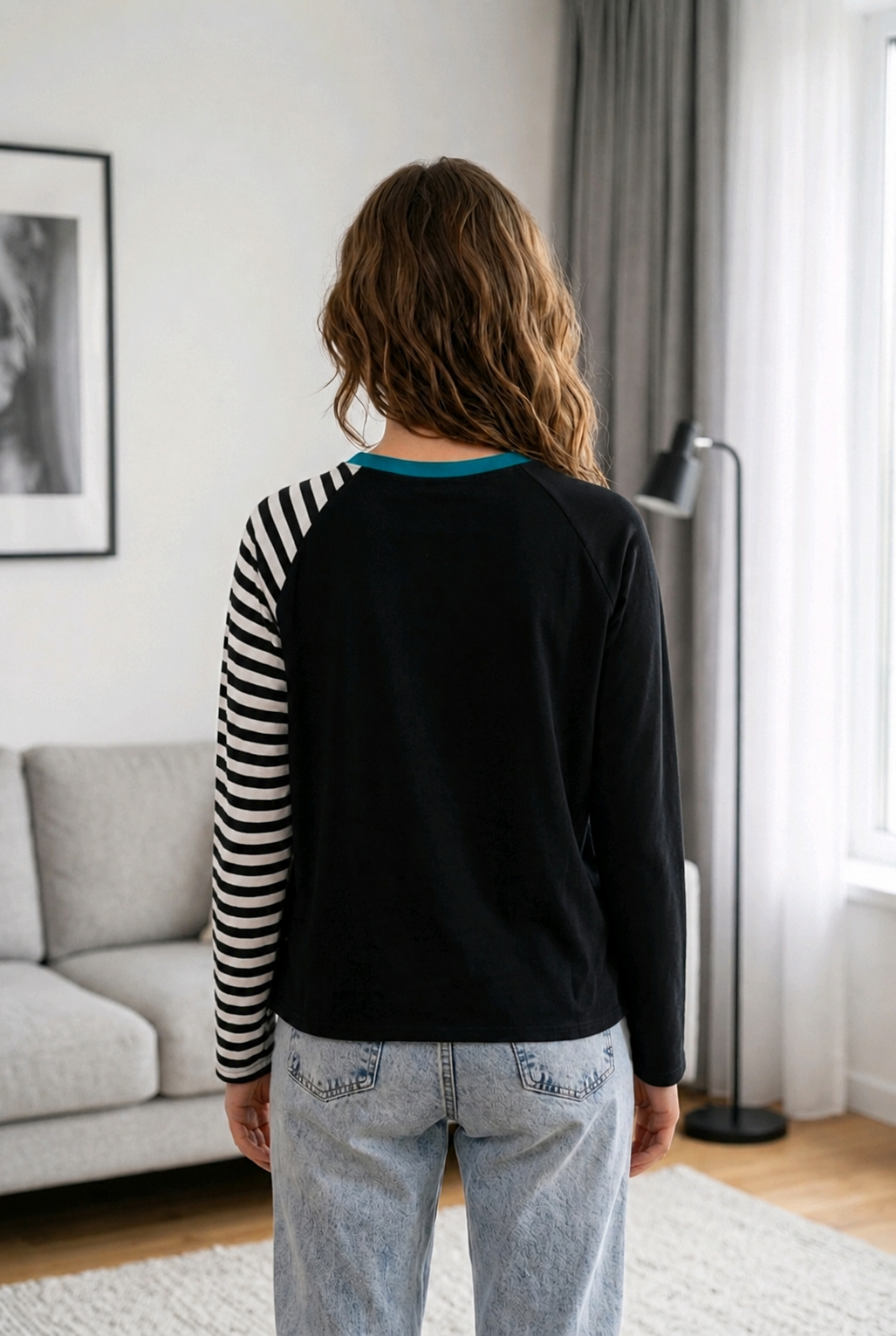 The back view of a woman standing in a living room, showcasing the solid black back panel of a modern raglan shirt. The asymmetrical design features one black and white striped sleeve and one solid black sleeve, with a teal neckband detail, paired with light-wash blue jeans as a DADI Design sewing pattern project.