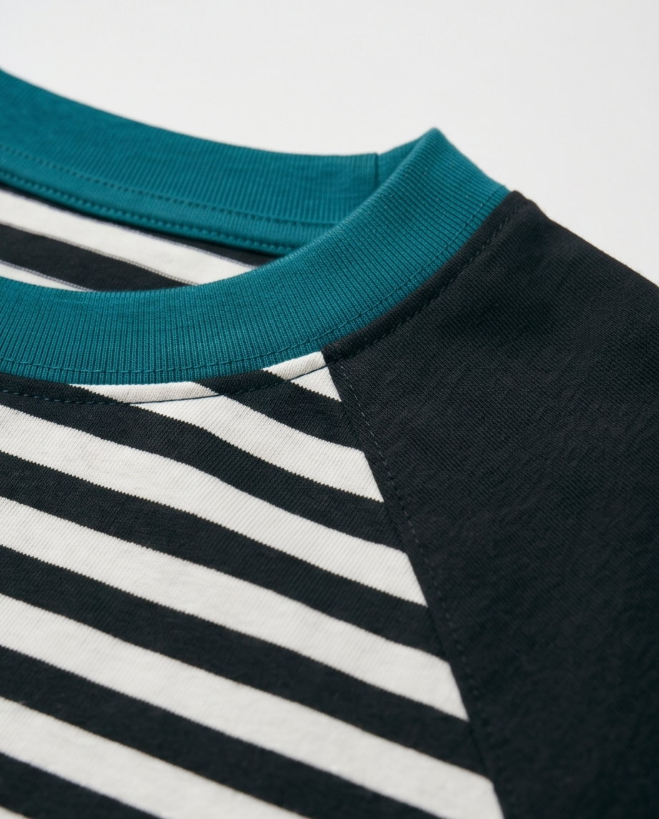 An extreme close-up view of a modern raglan shirt collar, showcasing a teal ribbed neckband precisely stitched onto a black and white striped knit front panel and a solid black raglan sleeve with clean topstitching details.