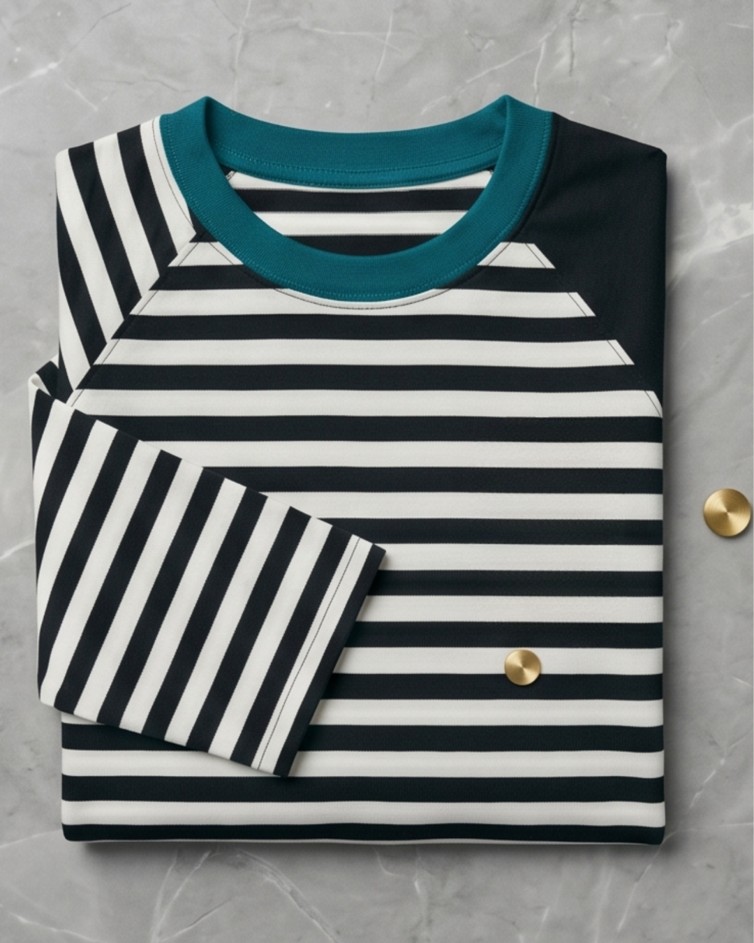A professional flat lay photograph of a neatly folded handmade raglan shirt by DADI Design, featuring a black and white striped front panel, a solid black raglan sleeve, and a contrasting teal neckband, beautifully arranged on a grey marble background.